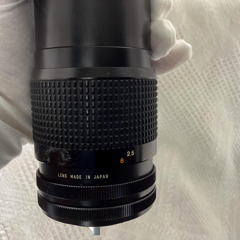 May include: Black camera lens with a ridged grip and a black barrel. The lens has white text that reads "2.5" and "8". The bottom of the lens reads "LENS MADE IN JAPAN".