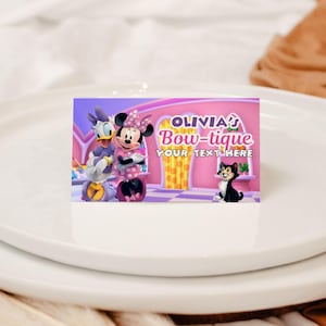 May include: A rectangular card featuring Minnie Mouse, Daisy Duck, and a black cat. The card has the text "OLIVIA'S Bow-tique" and "YOUR TEXT HERE" on a pink background. The card is on a white plate.