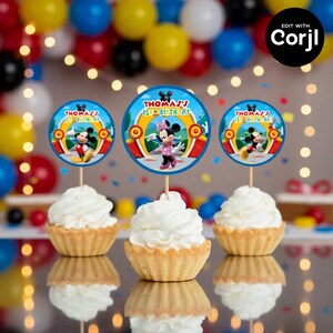 May include: Three cupcakes with white frosting, each topped with a circular decoration featuring Mickey and Minnie Mouse. The decorations read "Thomas' 1st Birthday" in blue and yellow. Colorful balloons and string lights are in the background.