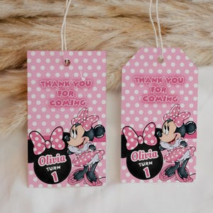 May include: Two pink gift tags with white polka dots and the text "Thank You For Coming." Each tag features a Minnie Mouse illustration and the name "Olivia" with "Turn 1." The tags have a string for hanging.