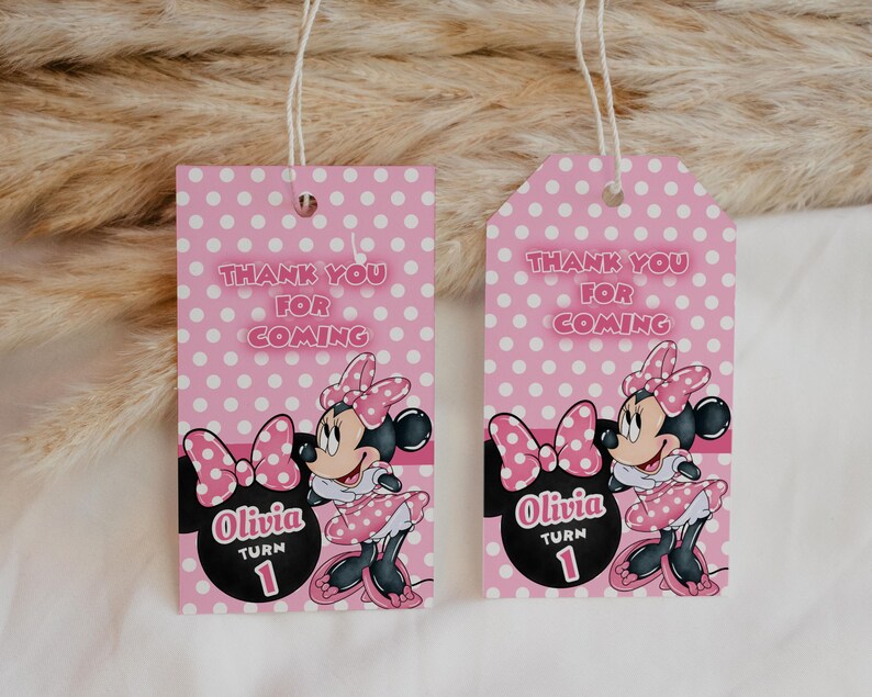 May include: Two pink gift tags with white polka dots and the text "Thank You For Coming." Each tag features a Minnie Mouse illustration and the name "Olivia" with "Turn 1." The tags have a string for hanging.