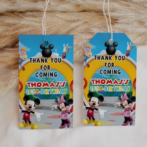 May include: Two birthday gift tags featuring Mickey and Minnie Mouse. The tags have a blue sky background with the text "Thank You For Coming" and "Thomas's 1st Birthday". The tags are rectangular and have a string attached.
