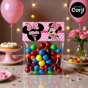May include: A clear cellophane bag filled with colorful candies, topped with a pink and white polka dot label featuring Minnie Mouse and the name "Olivia Turn 1". Pink balloons and a cake on a pink stand are in the background.