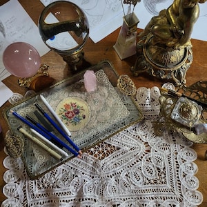 May include: A glass tray with a gold trim sits on a white lace doily. The tray contains several pencils, a pink crystal, and a small embroidered piece with flowers. The tray is on a wooden table with a gold cherub statue and a crystal ball.