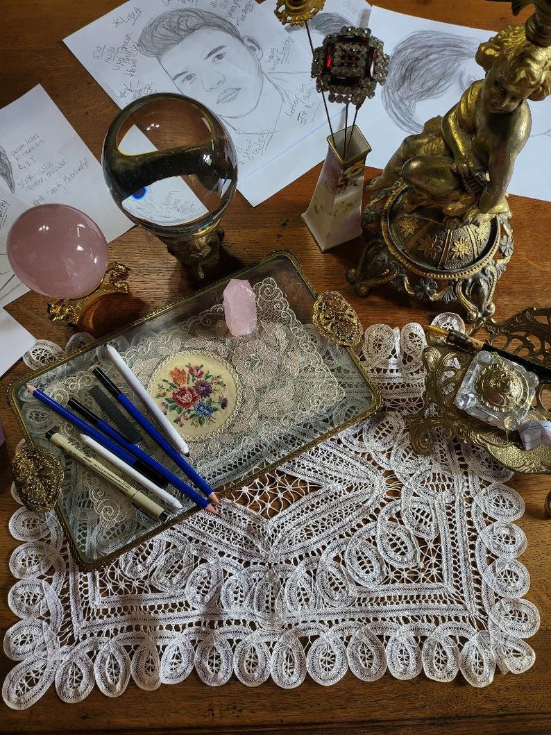 May include: A glass tray with a gold trim sits on a white lace doily. The tray contains several pencils, a pink crystal, and a small embroidered piece with flowers. The tray is on a wooden table with a gold cherub statue and a crystal ball.