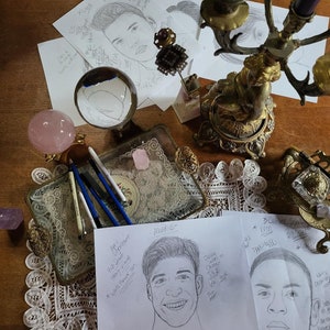 May include: A table with a white lace tablecloth and a variety of items, including a crystal ball, a pink crystal, a silver tray with pens, and three pieces of paper with hand-drawn portraits and astrological information. The paper with the text "AQUARIUS" has a portrait of a person with the text "Loyal, Independent, Free-Spirited, Friendly and Wants Freedom to Love". The paper with the text "CAPRICORN" has a portrait of a person with the text "Ruler Saturn in Capricorn, December 22 to January 19, Earth Sign, Practical, Responsible, Patient".