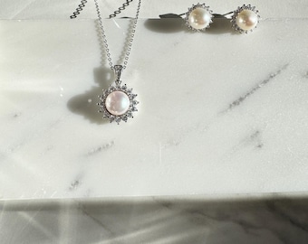 May include: A silver necklace with a pearl pendant surrounded by small clear stones. The necklace is paired with a set of pearl earrings with clear stones.