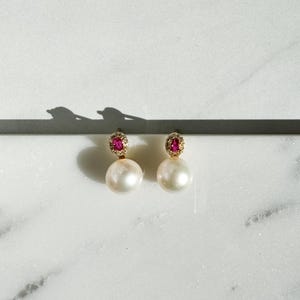 May include: A pair of elegant earrings featuring large, luminous white pearls. Each earring has a gold-colored top with a halo of small, clear stones surrounding a vibrant, round, red gemstone. The earrings are displayed on a white marble surface.