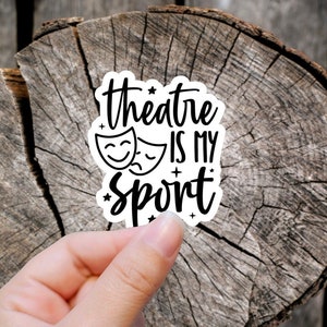 May include: White sticker with black text that reads "theatre is my sport" with two theatre masks on either side of the text. The sticker is on a wooden background.