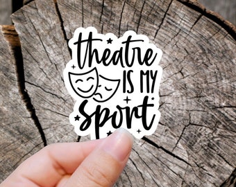 May include: White sticker with black text that reads "theatre is my sport" with two theatre masks on either side of the text. The sticker is on a wooden background.
