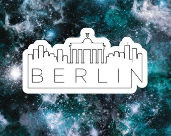 May include: A white sticker with a black outline of the Berlin skyline, including the Brandenburg Gate, and the word "BERLIN" underneath. The sticker is on a blue and black galaxy background.