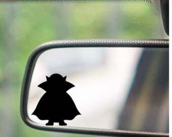 May include: A black silhouette of a character from a popular science fiction franchise is affixed to a car's rearview mirror. The character has large ears and a cloak. The background is a blurred view of the outside.