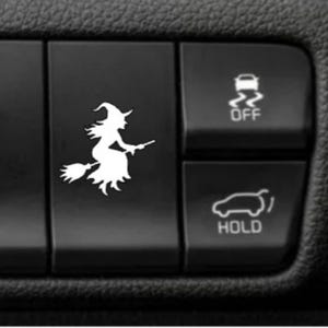 May include: A white vinyl decal of a witch flying on a broomstick applied to a black car interior button. The button is positioned next to two other black buttons featuring white icons for traction control off and a trunk hold function.