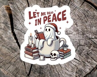 May include: A die-cut sticker featuring a ghost wearing a Santa hat, sitting in a large white mug while reading a book. The text reads LET ME READ IN PEACE. The scene includes stacks of books, two lit candles, a small cup, a skull, and a potted plant.