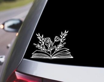 May include: A white vinyl decal of an open book with flowers growing out of it. The decal is on the back window of a silver car.