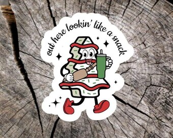 May include: A die-cut sticker featuring a cartoon character shaped like a triangular snack cake with white frosting, red stripes, and green sprinkles. The character has arms, legs, red shoes, and holds a green tumbler with the text out here lookin' like a snack.