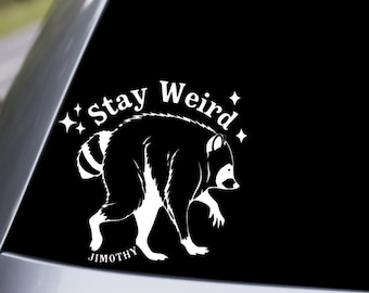May include: A white vinyl decal applied to a car window featuring a graphic of a raccoon in profile. The text "Stay Weird" is arched above the raccoon, accompanied by four-pointed star icons. The name "JIMOTHY" is printed in small text below.