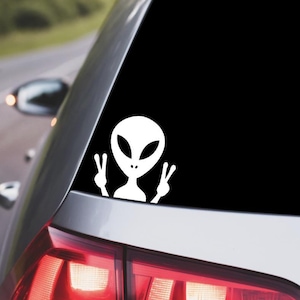 May include: White vinyl car decal featuring an alien with large oval eyes and a peace sign gesture. The decal is applied to the rear window of a silver car, with a road and trees visible in the background.