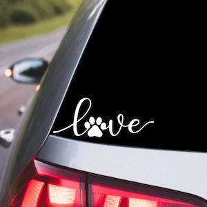 May include: A white vinyl decal with the word "love" written in a cursive font with a paw print in the middle of the word. The decal is on the back window of a silver car.