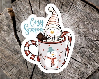 May include: A sticker featuring a snowman in a mug of hot chocolate with marshmallows and a candy cane. The mug has a snowman design and the words "Cozy Season" in a light blue font.