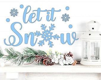 May include: A light blue wall decal featuring the text let it snow with a snowflake replacing the letter o. The design includes two additional small snowflakes and several blue dots. A white lantern and a frosted pine branch are nearby.
