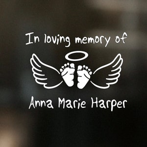 May include: White vinyl decal with the text "In loving memory of" above a pair of baby footprints with angel wings and a halo. Below the footprints is the text "Anna Marie Harper".