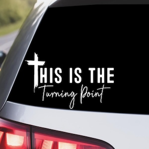 May include: White car decal with the text "THIS IS THE Turning Point" in a script font. The decal features a white cross graphic. The image is taken from a slightly angled perspective, showing the car's rear window.