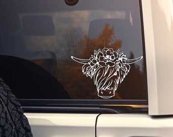 May include: White vinyl decal of a Highland cow with floral accents, applied to the rear window of a white SUV. The design features the cow's head with long horns and a floral crown.