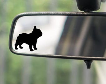 May include: A black silhouette of a French Bulldog dog is affixed to a car's rearview mirror. The dog is facing left, with its distinctive features clearly visible. The mirror is framed in silver, reflecting a blurred outdoor scene.