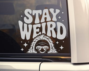 May include: A white vinyl decal applied to a vehicle window featuring the text STAY WEIRD in a bold, wavy font. Below the text is a detailed graphic illustration of a furry creature resembling a Bigfoot, surrounded by small four-pointed stars.