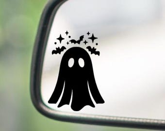 May include: A black vinyl decal applied to a vehicle side mirror. The design features a solid black ghost silhouette with two oval eyes, surrounded by small flying bats and four-pointed star shapes arranged in a semi-circle above the head.