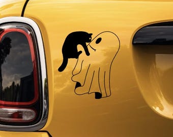 May include: A black vinyl decal applied to a yellow surface, featuring a silhouette of a black cat climbing onto the back of a simple, line-drawn ghost character. The ghost has a single oval eye and a small, curved line representing a mouth.