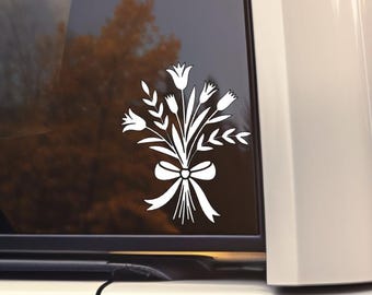 May include: White floral bouquet decal with a bow, applied to a car window. The design features stylized flowers and leaves, creating a decorative accent. The decal is a solid white color, contrasting against the dark window.
