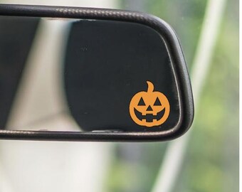 May include: A close-up of a car's rearview mirror with a small orange jack-o'-lantern sticker. The sticker has a carved face with triangular eyes and a smiling mouth. The mirror is black and has a dark frame.