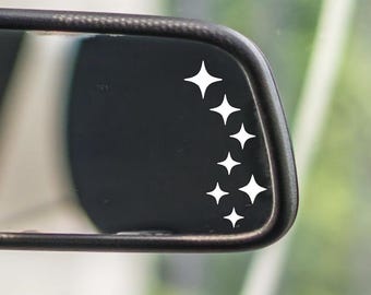 May include: A black car rearview mirror with a series of white starburst decals. The mirror is set against a blurred background, suggesting an outdoor setting. The decals are arranged in a descending diagonal line.