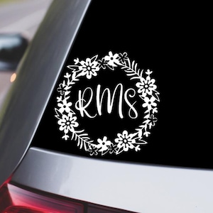 May include: A white vinyl decal applied to a car window, featuring a circular floral wreath design surrounding the letters RMS in a cursive script font. The floral elements include various stylized blossoms, leaves, and stems in a solid white color.