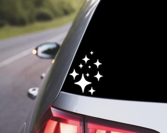 May include: White starburst decal applied to the rear window of a silver car. The decal features a cluster of various-sized, white star shapes. The car is parked on a road with a blurred green background.