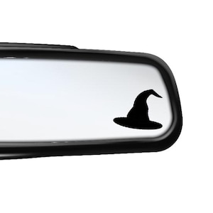 May include: A black rear-view mirror with a black witch hat sticker on the reflective surface. The witch hat is a silhouette, with a wide brim and a pointed top. The mirror is set against a white background.