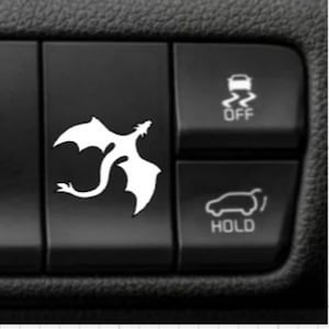 May include: A black rectangular car interior button featuring a white silhouette of a flying dragon. To the right, two additional black buttons are visible, one displaying a car icon with the text OFF and another with a car icon and the text HOLD.