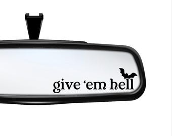 May include: A black car rearview mirror with a white decal applied to the glass. The decal features the black text 'give 'em hell' in a serif font, accompanied by a small black silhouette of a flying bat positioned above the word hell.