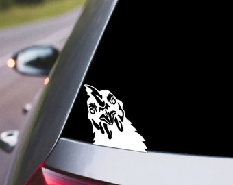 May include: A white vinyl decal of a chicken peeking out, applied to a car window. The chicken's head is in a stylized, cartoonish design with black details. The decal is on the rear window of a silver car.