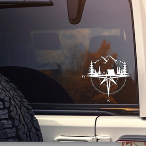 May include: White vinyl decal of a compass with mountains, trees, and a tent inside. The compass points are labeled with the letters N, S, E, and W.