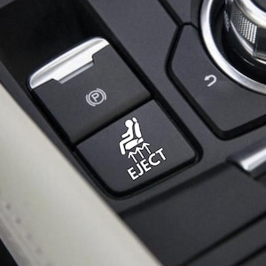 May include: Close-up of a black car console with several buttons. The image highlights a button labeled "EJECT" with a graphic of a person in a seat. Other buttons include a parking brake symbol and a curved arrow.