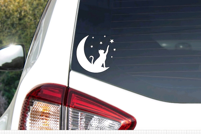 May include: White vinyl car decal of a cat reaching for a star above a crescent moon. The decal is on a black car window.