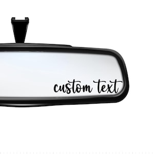 May include: A black rearview mirror with a white background and the text "custom text" in black cursive lettering.