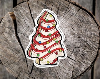 May include: A Christmas tree sticker with a white base, red ribbon, and colorful star and dot ornaments. The sticker is on a wooden surface.