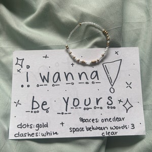May include: A white card with the text "I wanna be yours" written in black marker. The text is separated by dashes and dots. There are gold and white beads on the card, forming a circle. The card also includes the text "dots: gold, dashes: white, spaces: oneclear, space between words: 3 clear".