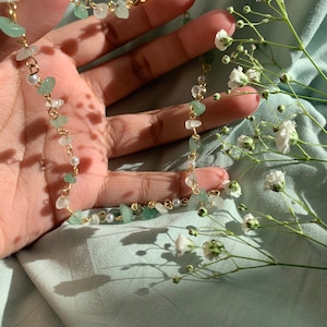 May include: A delicate gold chain necklace with small green and white stones and pearl accents. The necklace is displayed on a light blue fabric background with white flowers.