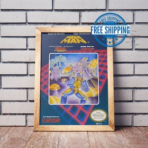 May include: A framed print of a vintage video game cartridge box art for Mega Man. The box art features a blue robot character with a yellow helmet and a red background. The text on the box art reads "Mega Man" and "Game Pak by Capcom".