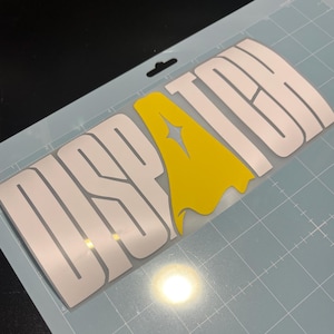 May include: A white vinyl decal with the word "DISPATCH" in a stylized font, with a yellow graphic in the center. The decal is on a light blue cutting mat.